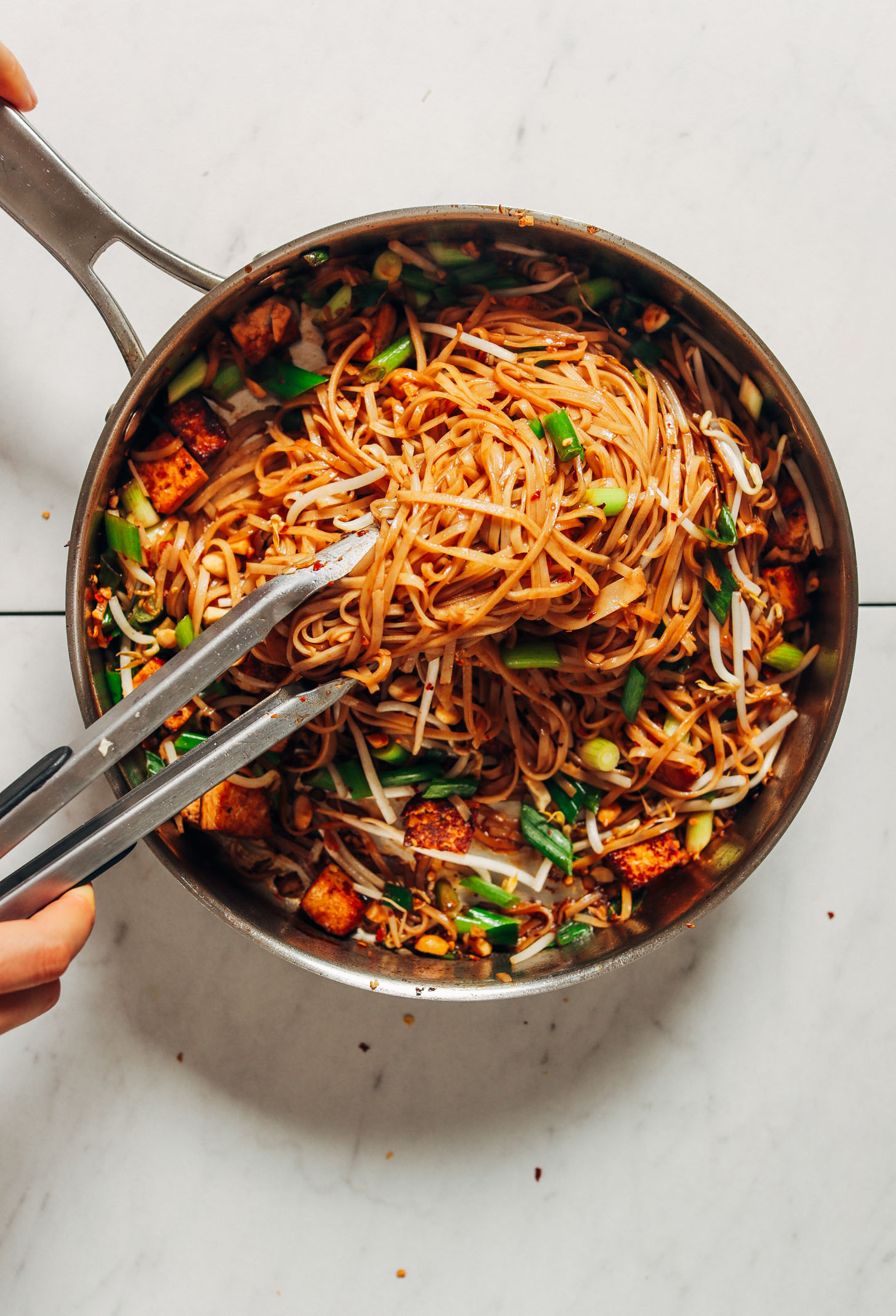 Using tongs to toss pad thai noodles with sauce, green onions, and bean sprouts