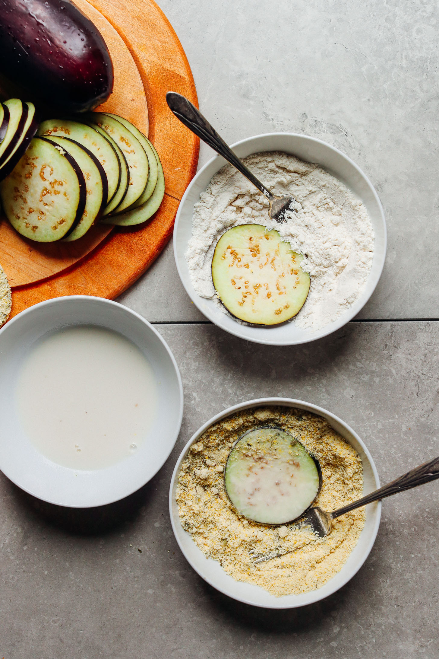 Sliced eggplant with bowls of almond milk, gluten-free flour, and dry coating for breading the CRISPY Eggplant Parmesan