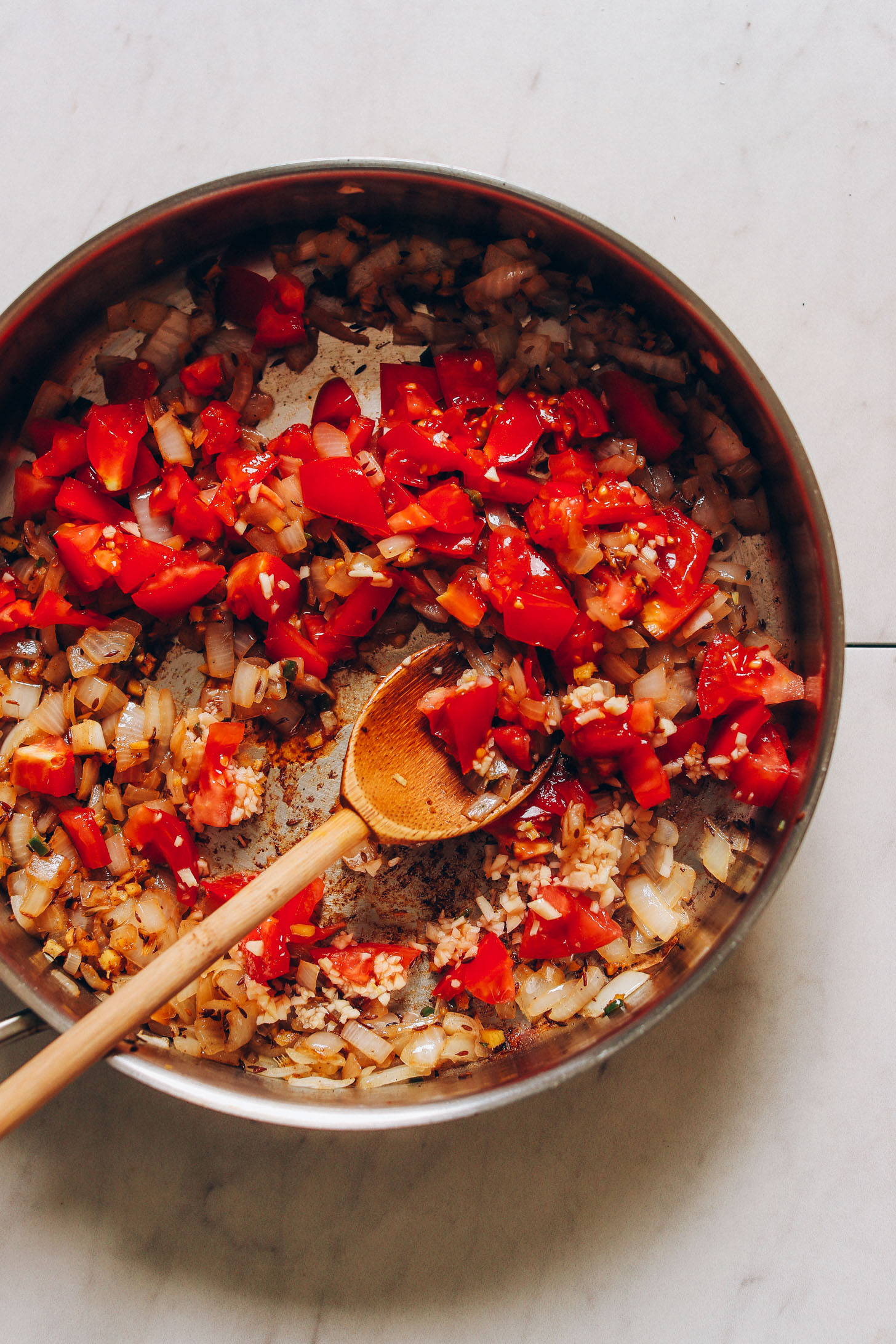 Overhead image of tomatoes, onion, and garlic being sautéed in a skillet with a wooden spoon stirring the ingredients