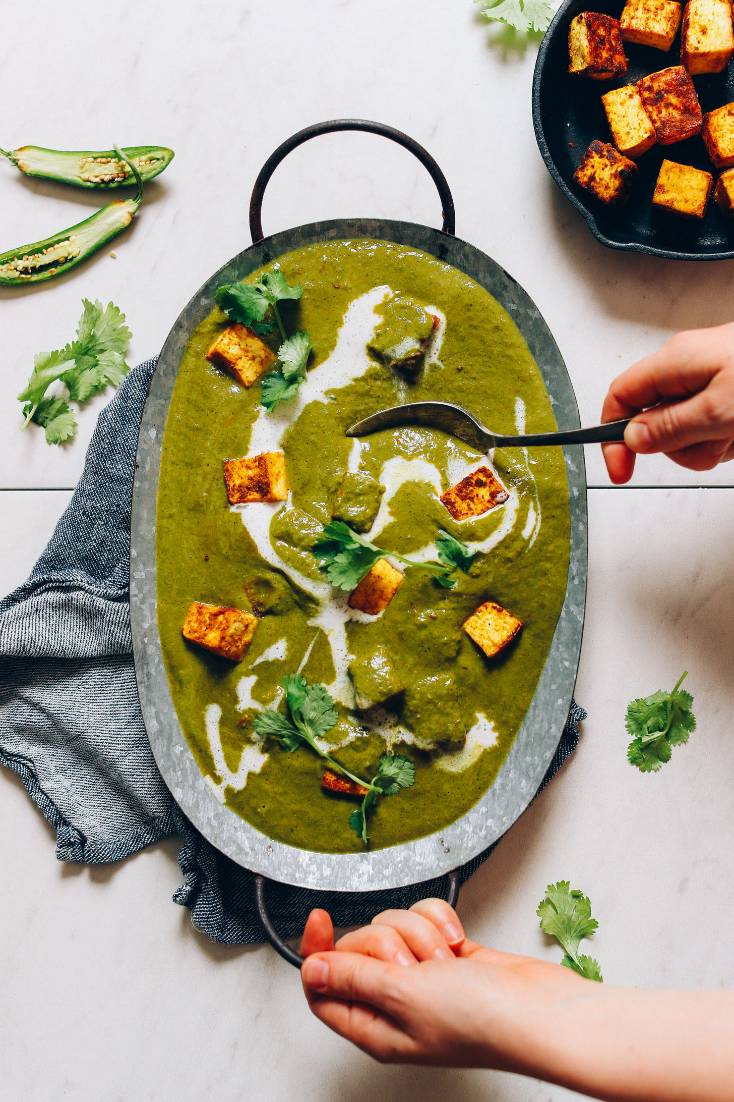 Overhead image of palak paneer with tofu in a metal tin and a hand with a spoon scooping up a serving