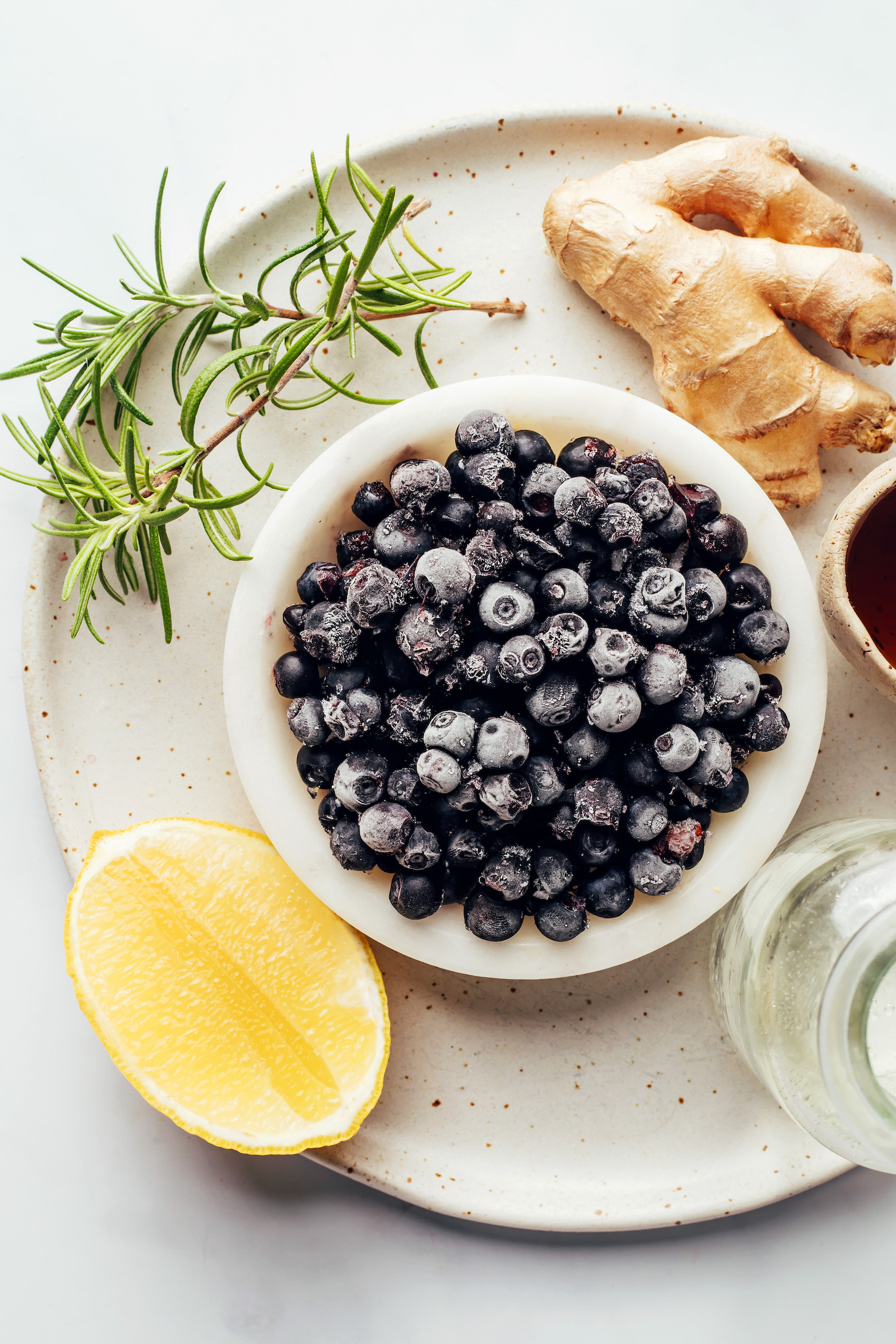 A plate with frozen blueberries, fresh rosemary, fresh ginger, lemon, and maple syrup