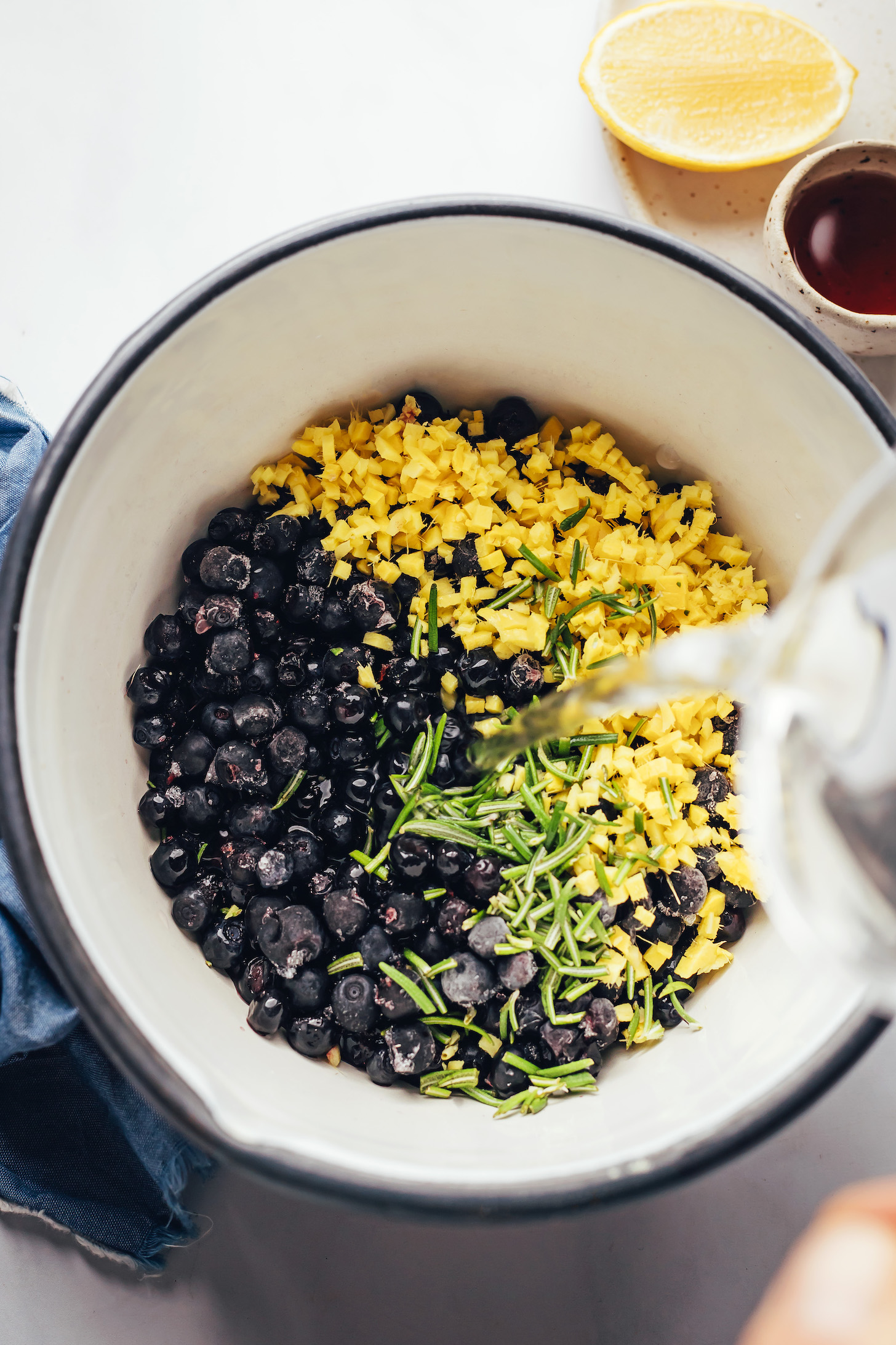 Water being poured into a saucepan with blueberries, fresh ginger, and fresh rosemary