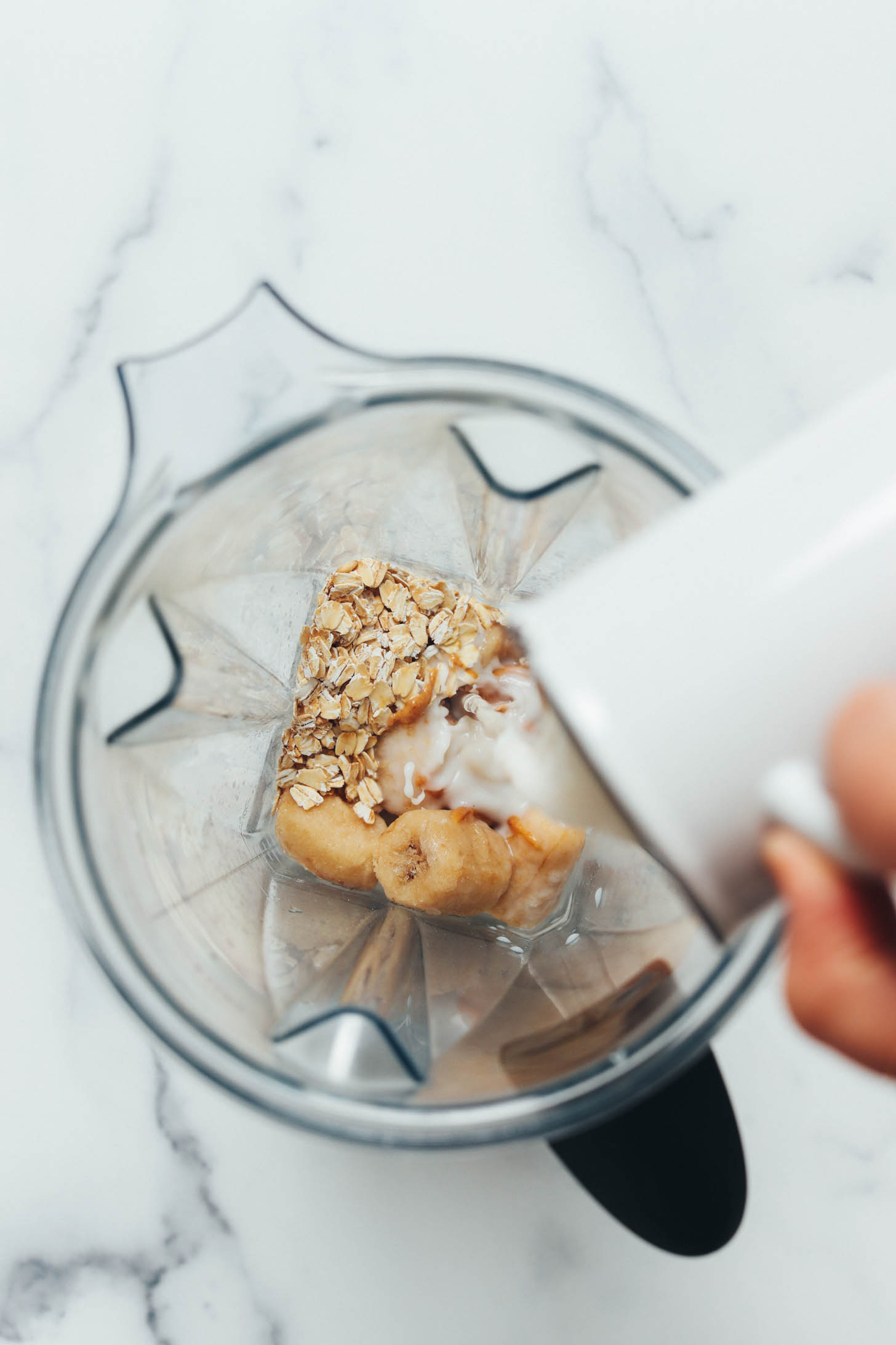 Pouring almond milk over frozen banana and rolled oats in a blender