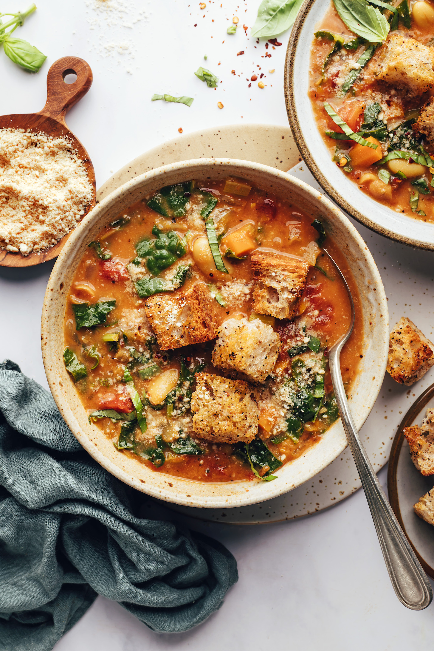 Small plates of vegan parmesan and croutons next to and on bowls of Tuscan white bean and kale soup