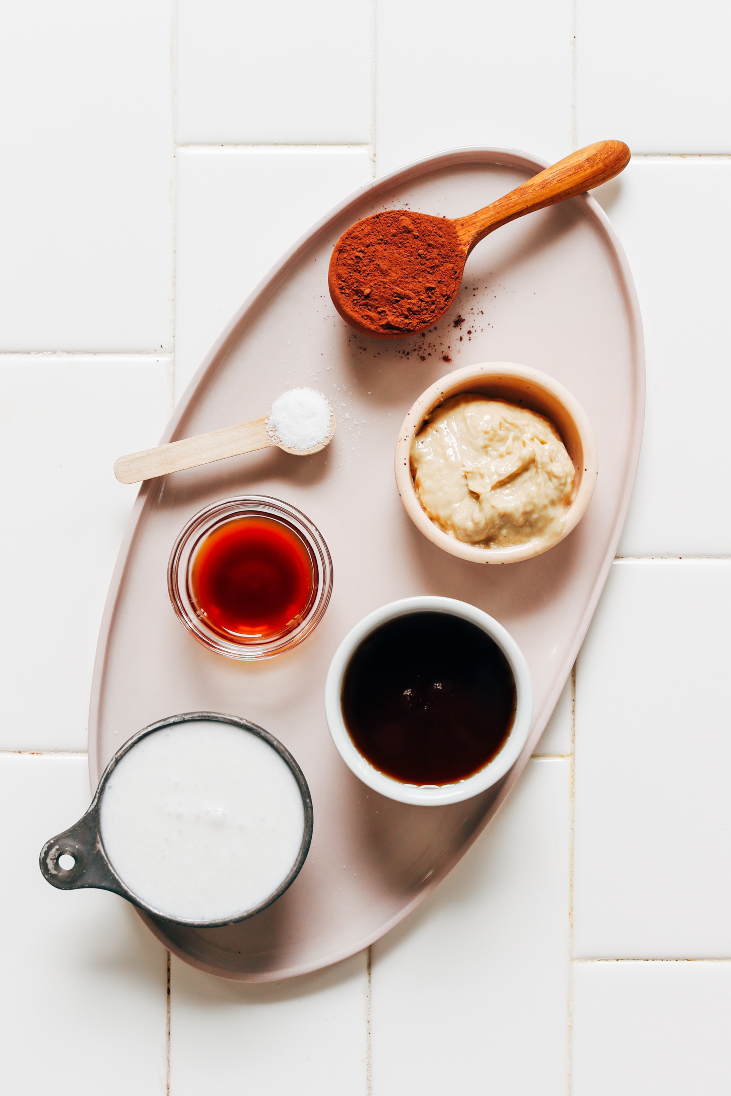 Tray with spoonfuls and bowls of cocoa powder, cashew butter, maple syrup, coconut milk, vanilla extract, and sea salt