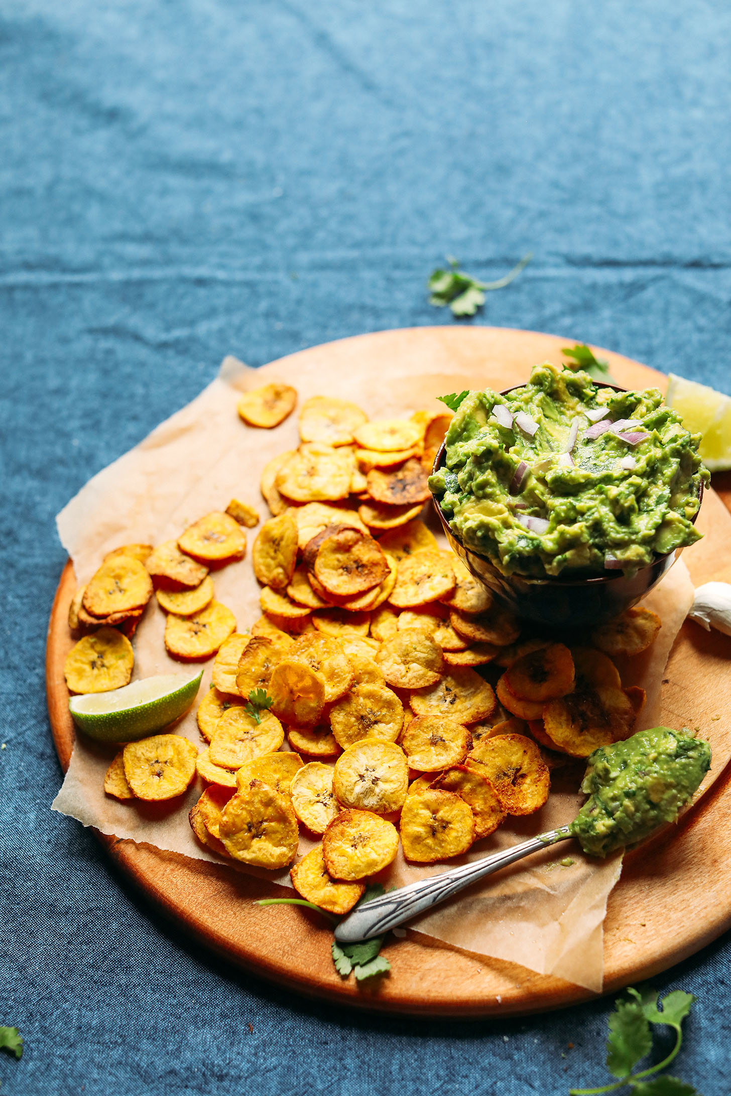 Wood cutting board with homemade Baked Plantain Chips and a bowl of Garlicky Guacamole