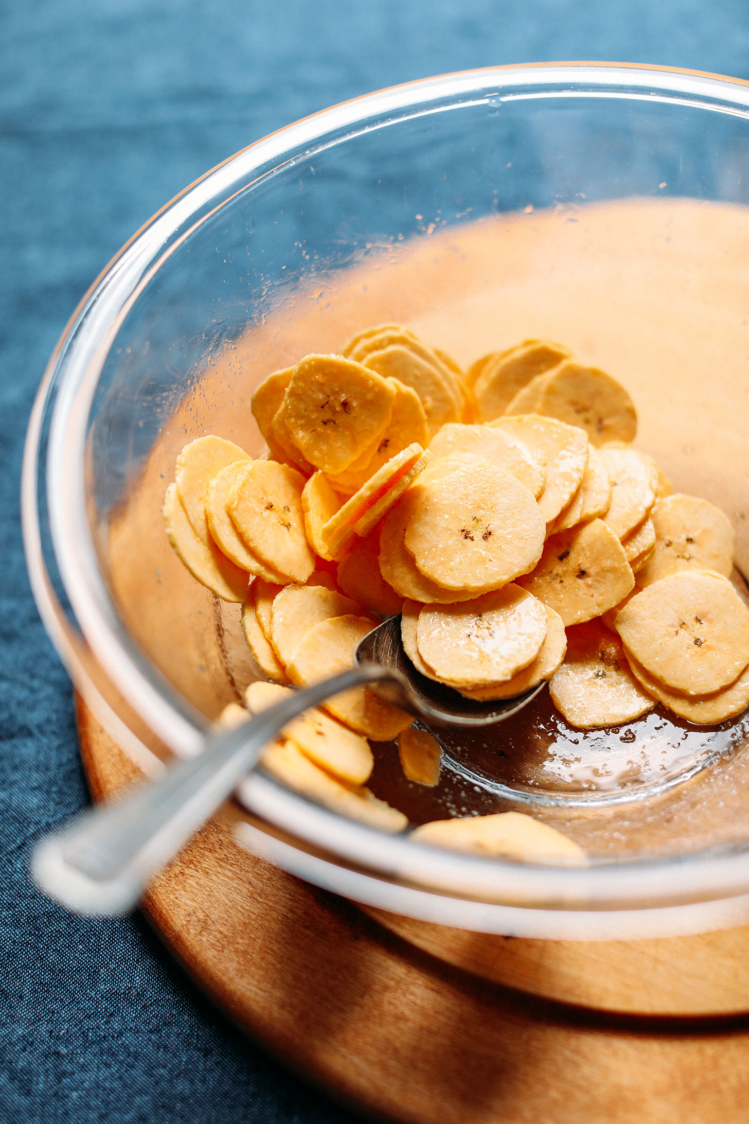 Stirring thinly sliced plantains in a bowl with melted coconut oil and salt