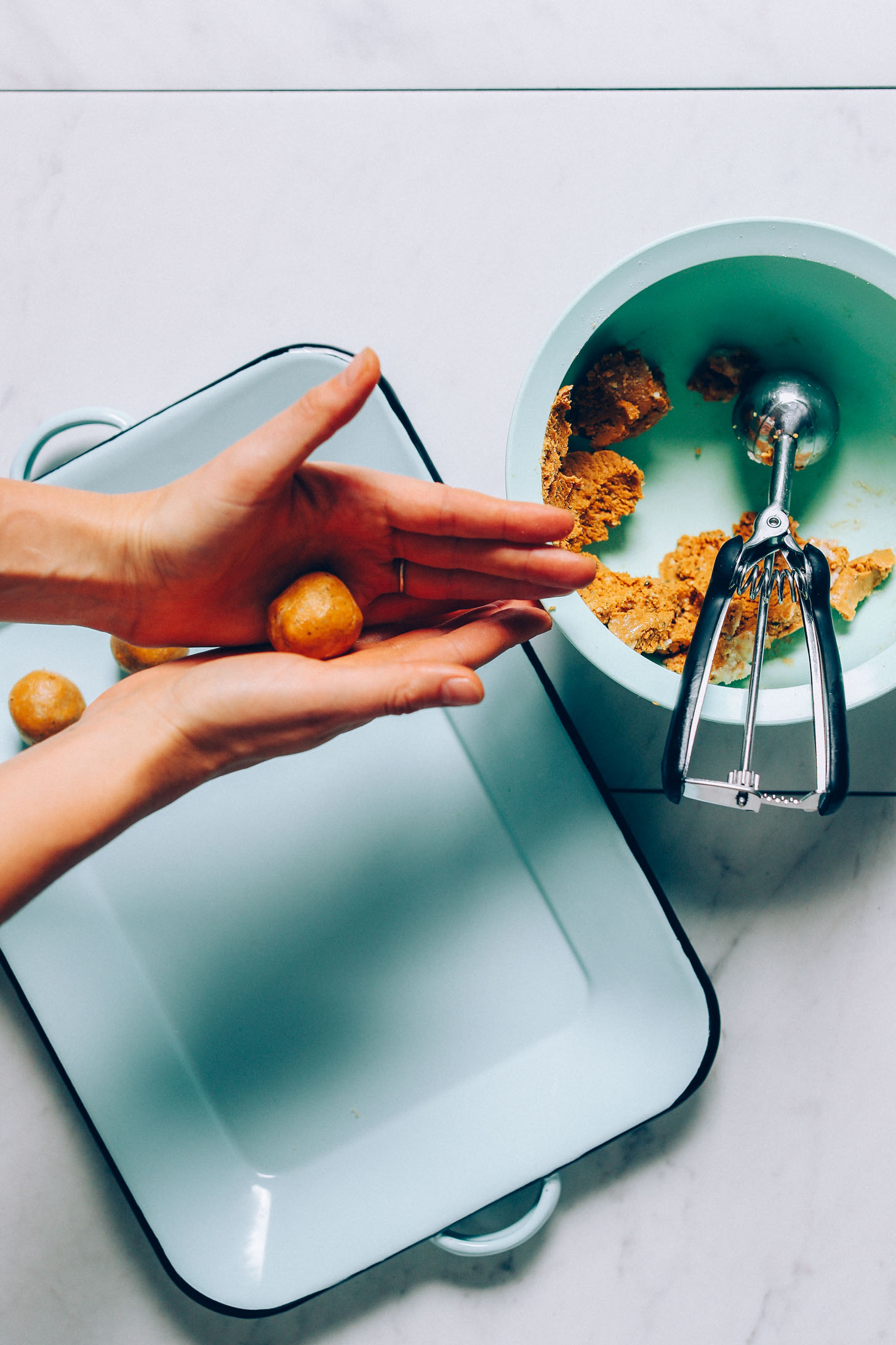 An overhead image of golden milk snack bites being rolled into bite size balls with a blue tray underneath and mixing bowl to the right with cookie scoop