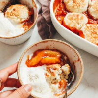 Bowl of gluten-free peach cobbler with a scoop of vegan vanilla ice cream next to a baking dish with more cobbler