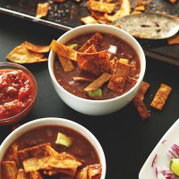 Two bowls of Loaded Veggie Nacho Soup beside bowls of salsa and chopped red onion and lime