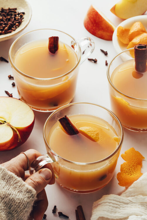 Mugs of hot spiced apple cider with a hand holding one mug