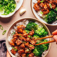 Using chopsticks to pick up a bite of sesame tofu from a bowl with rice and steamed broccoli